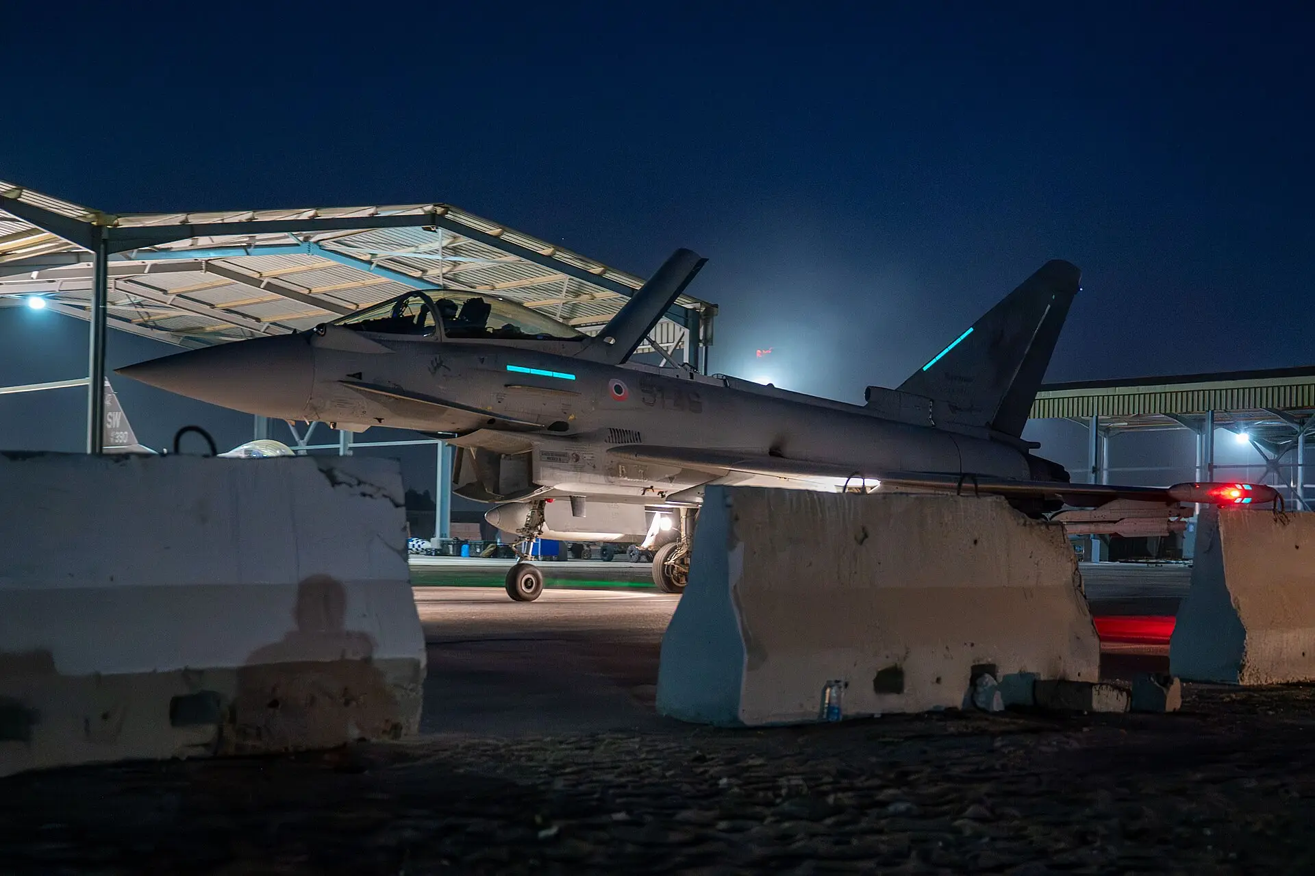 A combat aircraft parked under floodlights on an airfield at night.